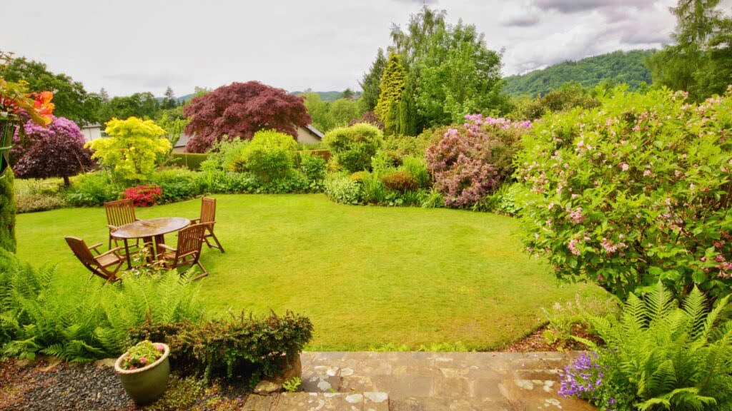 widok z tarasu na ogród, na środku trawnik ze stołem i krzesłami, dookoła różnobarwne nasadzenia