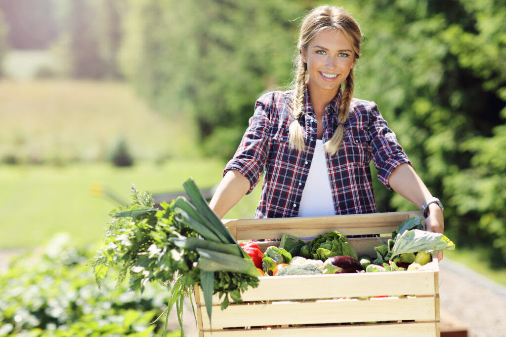 Kobieta w warkoczach i koszuli w kratkę niesie skrzynkę z warzywami.