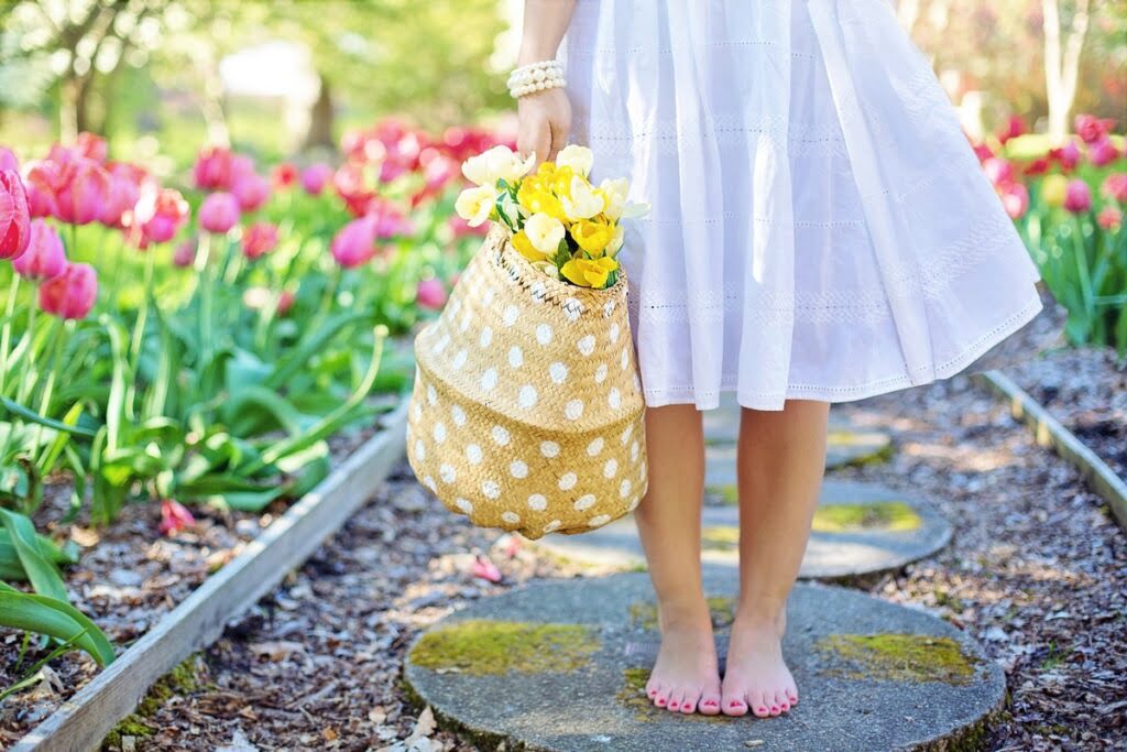 Dziewczyna w białej sukience z torbą z tulipanami, stojąca na ścieżce przy rabacie tulipanowej.